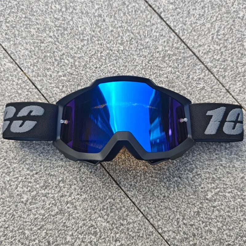 Une paire de lunettes de moto tout-terrain noires avec des verres réfléchissants bleu vif posées sur une surface grise.