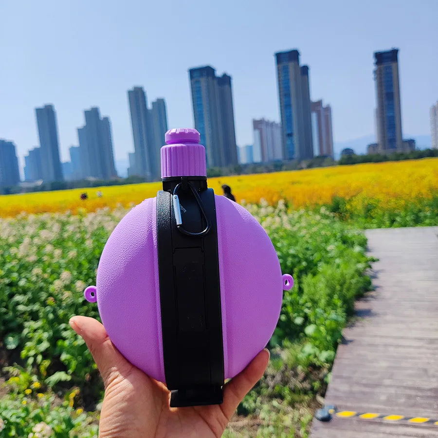 A hand holds a purple collapsible silicone camping kettle outdoors with a city skyline in the background.