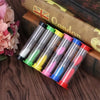 A set of six colorful glass sand timer hourglasses arranged on a wooden surface next to a book.