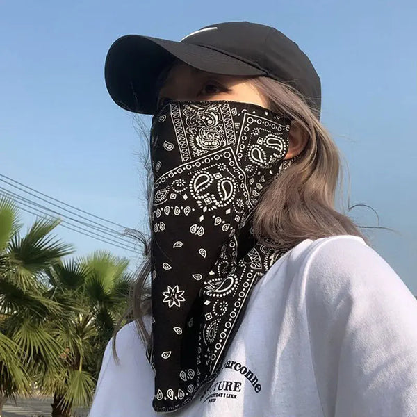 Eine Frau trägt einen schwarzen Paisley-UV-Schutz-Halstuch und eine Gesichtsmaske unter einem schwarzen Hut, im Freien.