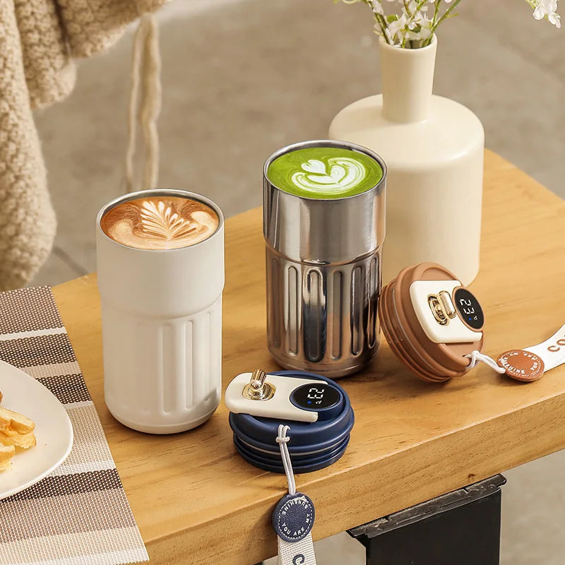 A stainless steel insulated thermos bottle next to a white ceramic mug and a glass vase on a wooden table.