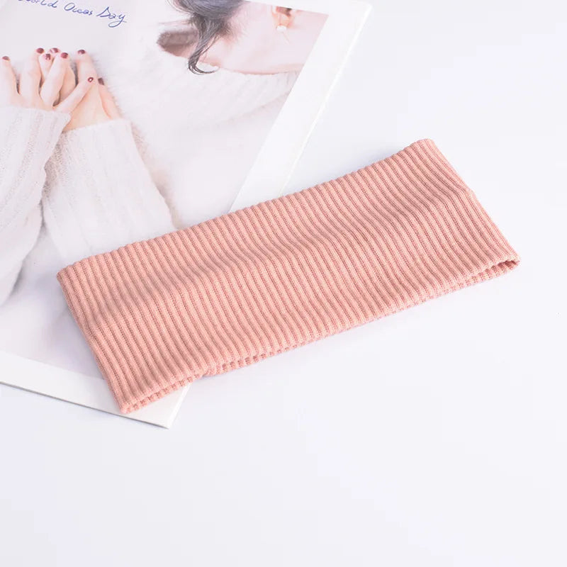 A pink ribbed knitted headband is displayed next to a book and hands.