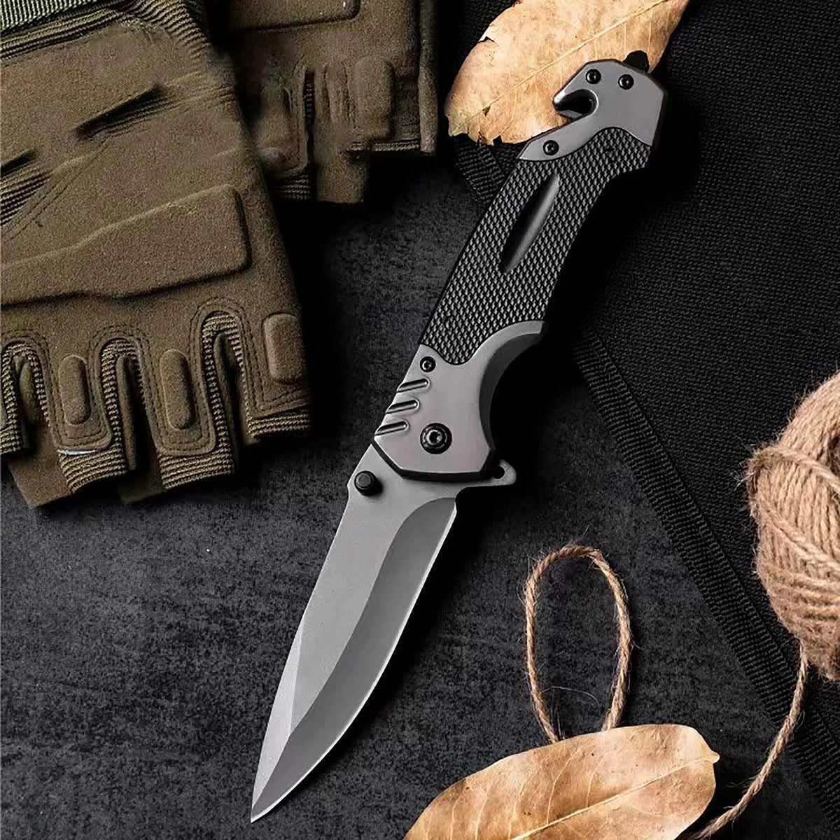 A multi-functional folding survival knife is shown next to a tactical glove and dried leaves on a dark surface.