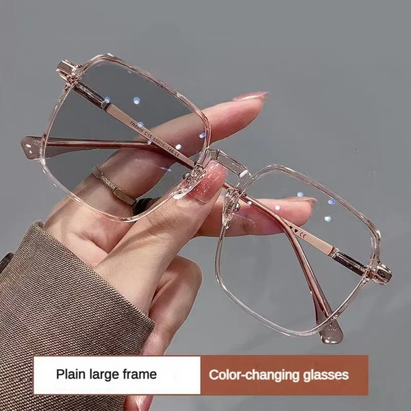 A hand holds a pair of clear, rose gold-toned photochromic blue light blocking glasses against a gray background.