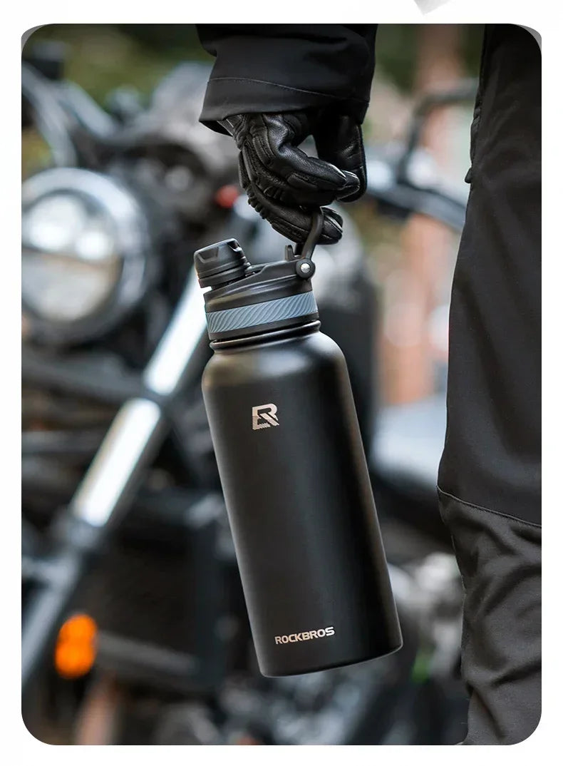 A black insulated stainless steel thermal water bottle is being held by a gloved hand in an outdoor setting.