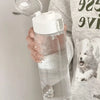 A hand holds a large capacity transparent sports water bottle with a clear lid against a light gray t-shirt.