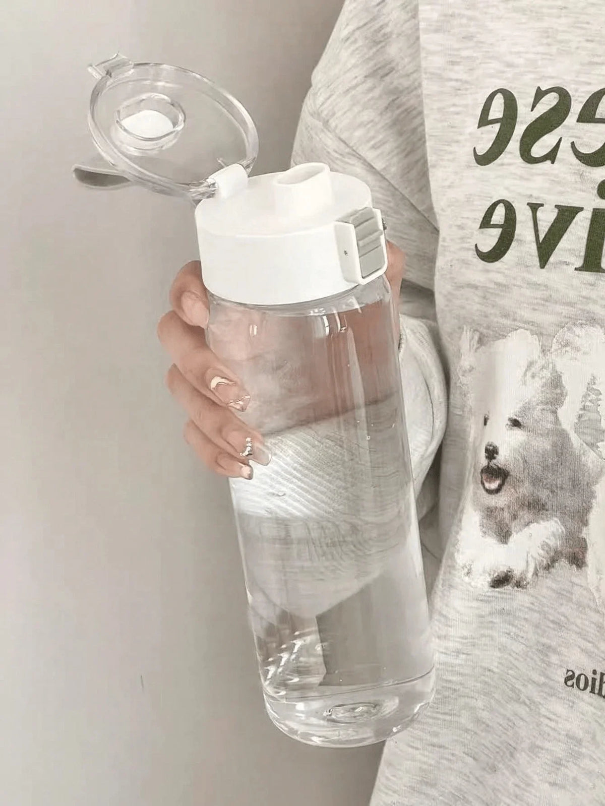 A hand holds a large capacity transparent sports water bottle with a clear lid against a light gray t-shirt.