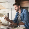 A man in a denim shirt is using a laptop and a smartphone in a modern indoor setting.