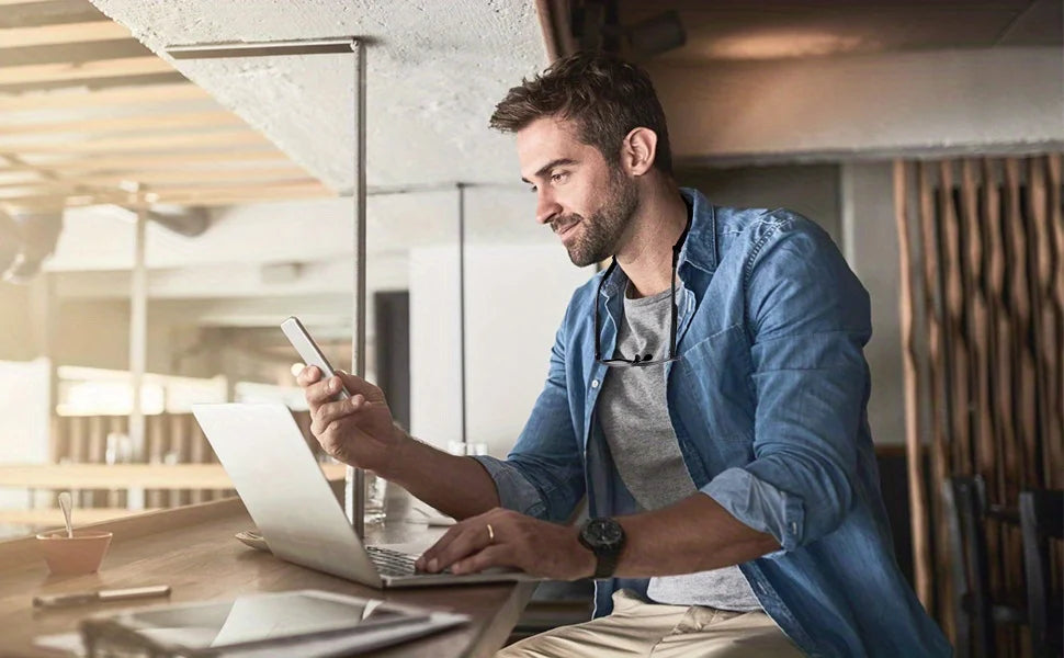 A man in a denim shirt is using a laptop and a smartphone in a modern indoor setting.