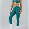A woman wearing teal high-waist yoga leggings and a matching sports bra, standing against a light gray background.
