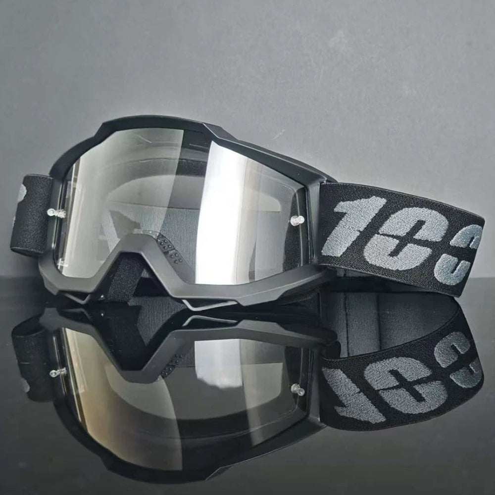 Un par de gafas de motocross negras con lentes transparentes y una correa negra, mostradas sobre una superficie reflectante.