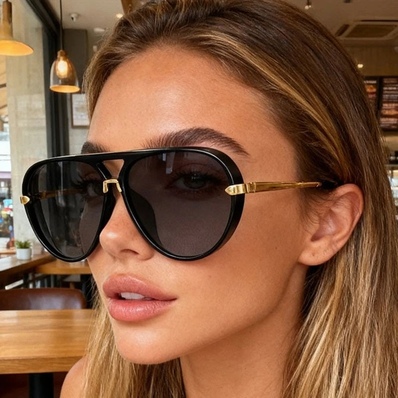 A close-up of a woman wearing classic oval pilot sunglasses with gold accents against a blurred indoor background.