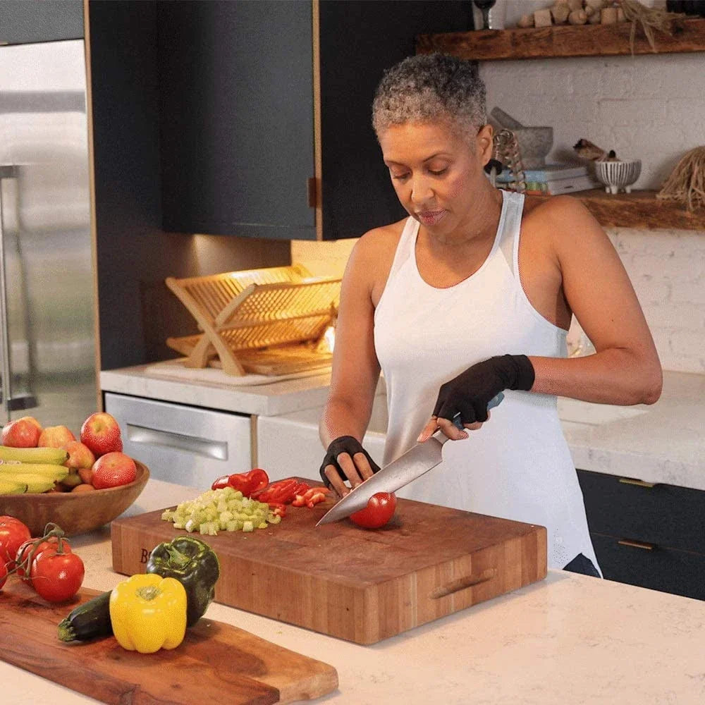 A woman in a white tank top is using a knife to cut vegetables on a wooden cutting board.