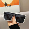 A hand holds a pair of black and white oversized square sunglasses against an orange background.