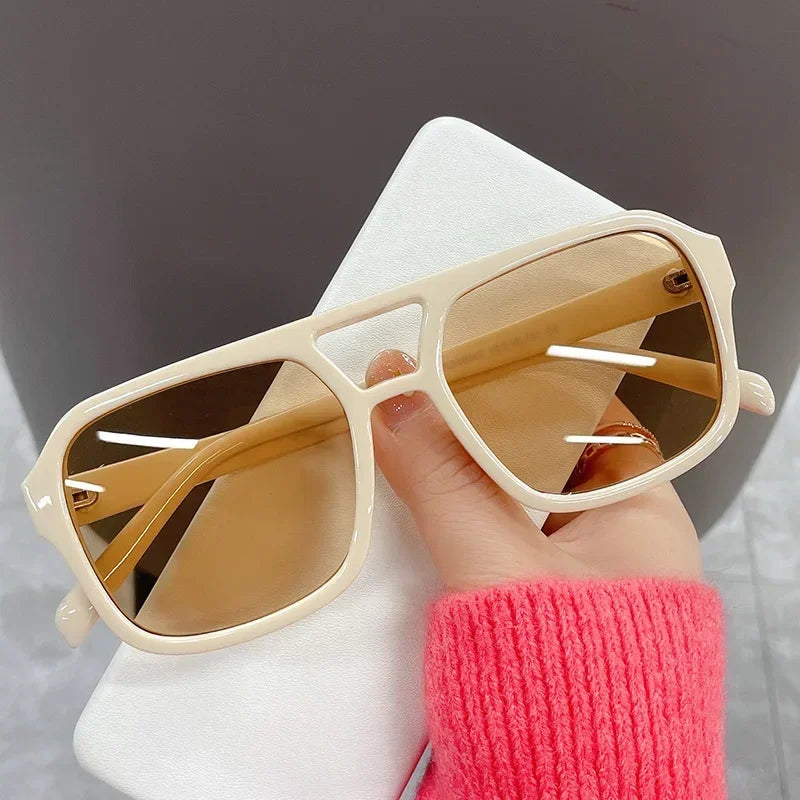 Una mano sostiene unas gafas de sol cuadradas beige claro con monturas de metal fino contra un fondo blanco.