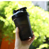 A hand holds a black leak-proof shaker bottle against a blurred green outdoor background.