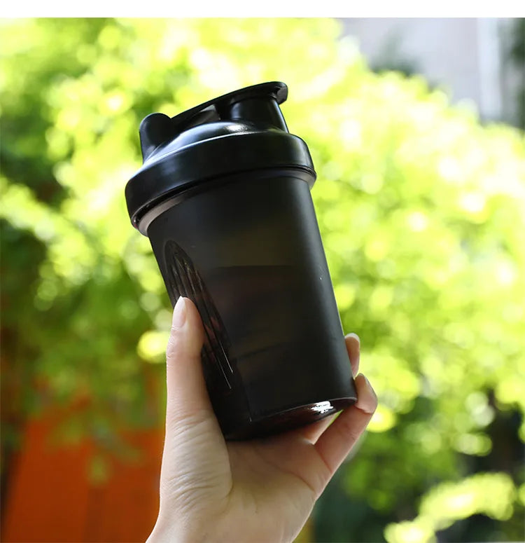 A hand holds a black leak-proof shaker bottle against a blurred green outdoor background.