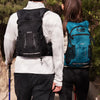A woman wearing a white shirt and black pants stands with a hydration backpack on her back in a wooded area.
