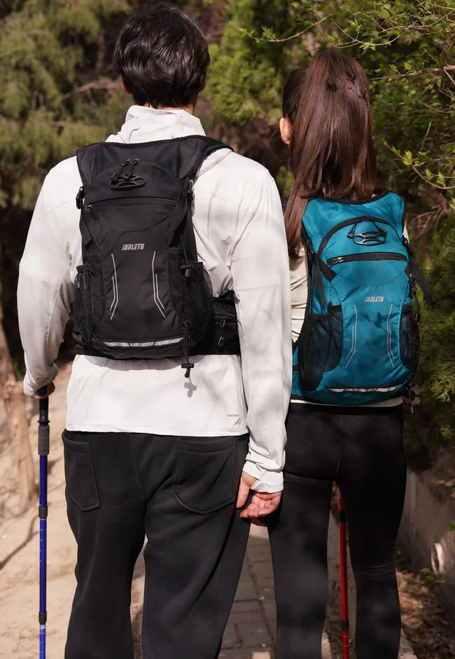 A woman wearing a white shirt and black pants stands with a hydration backpack on her back in a wooded area.