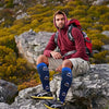 A man wearing a maroon hoodie and khaki pants sits on rocks outdoors, modeling women's athletic knee-high compression socks.