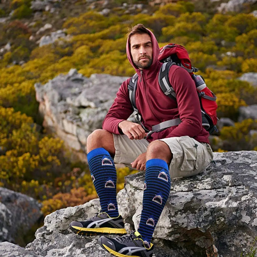 A man wearing a maroon hoodie and khaki pants sits on rocks outdoors, modeling women's athletic knee-high compression socks.