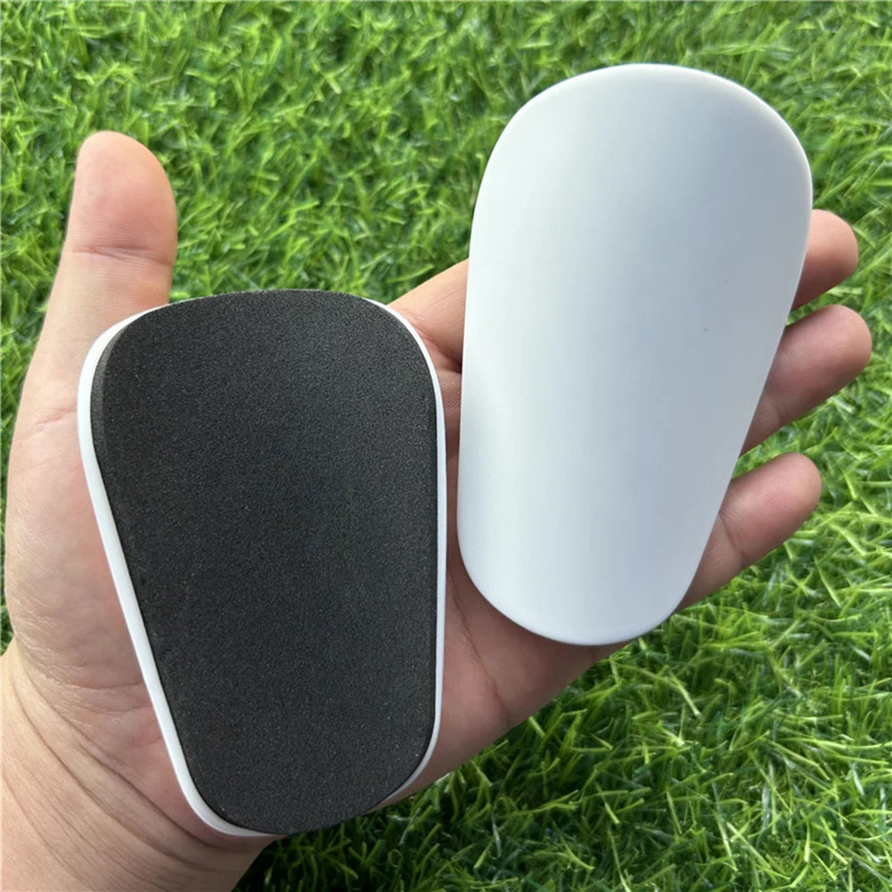 Una mano sostiene una protectora de espinilla mini blanca y negra sobre césped verde.
