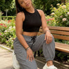A woman wearing black crop top and gray drawstring cargo joggers sits on a wooden bench outdoors.