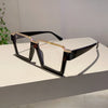 A pair of oversized square semi-rim metal eyeglasses with dark frames and gold accents resting on a white surface.