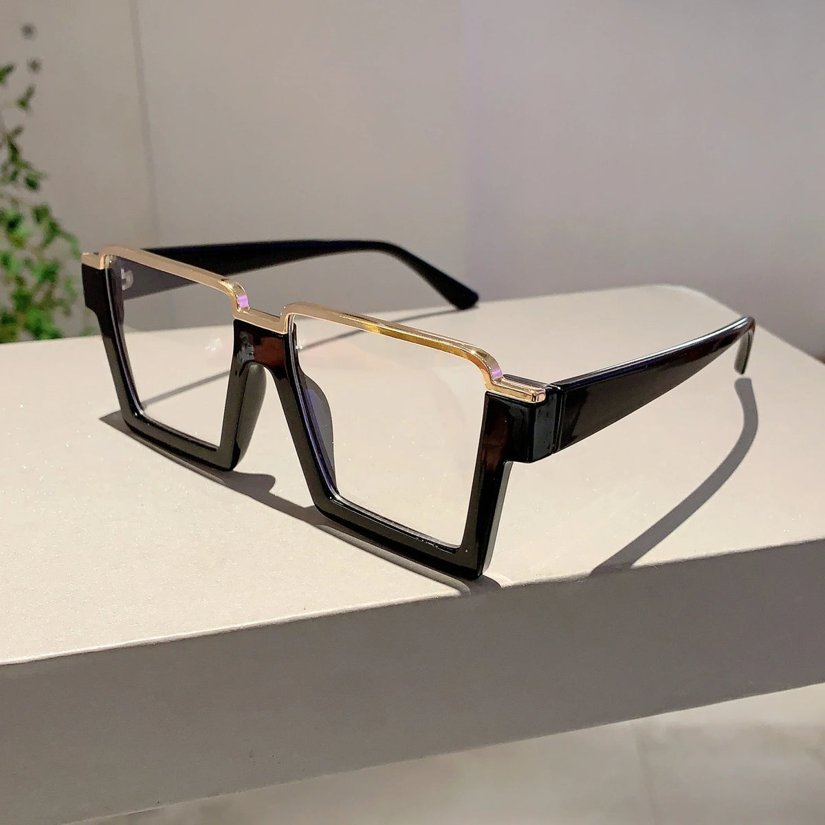 A pair of oversized square semi-rim metal eyeglasses with dark frames and gold accents resting on a white surface.