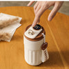 A hand is holding a white insulated thermos bottle with a black smart display and a leather cord tag on a wooden table.