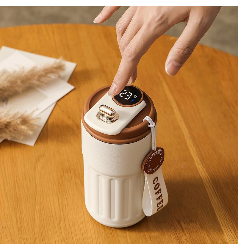 A hand is holding a white insulated thermos bottle with a black smart display and a leather cord tag on a wooden table.