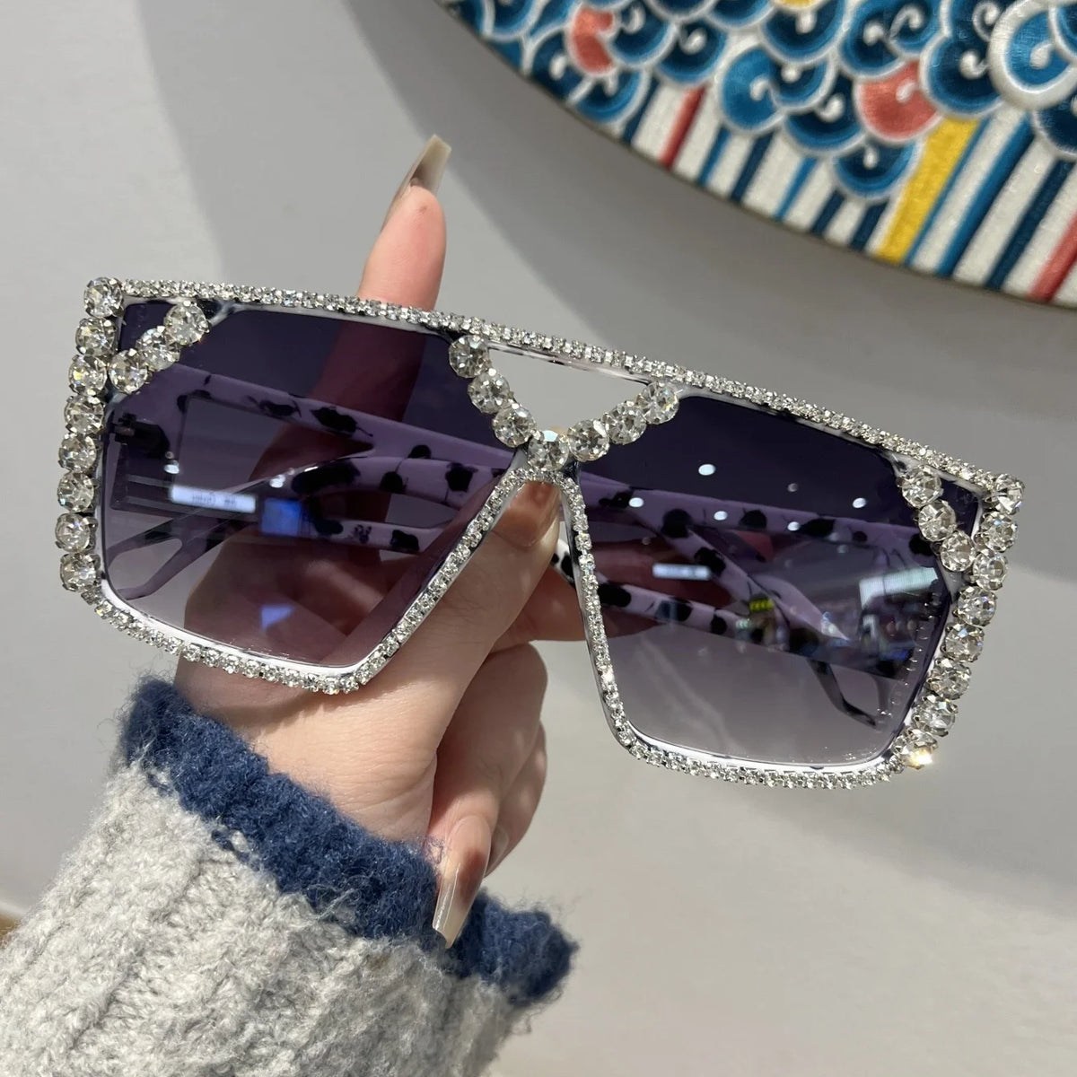 A hand holds a pair of geometric rhinestone fashion sunglasses against a colorful patterned background.