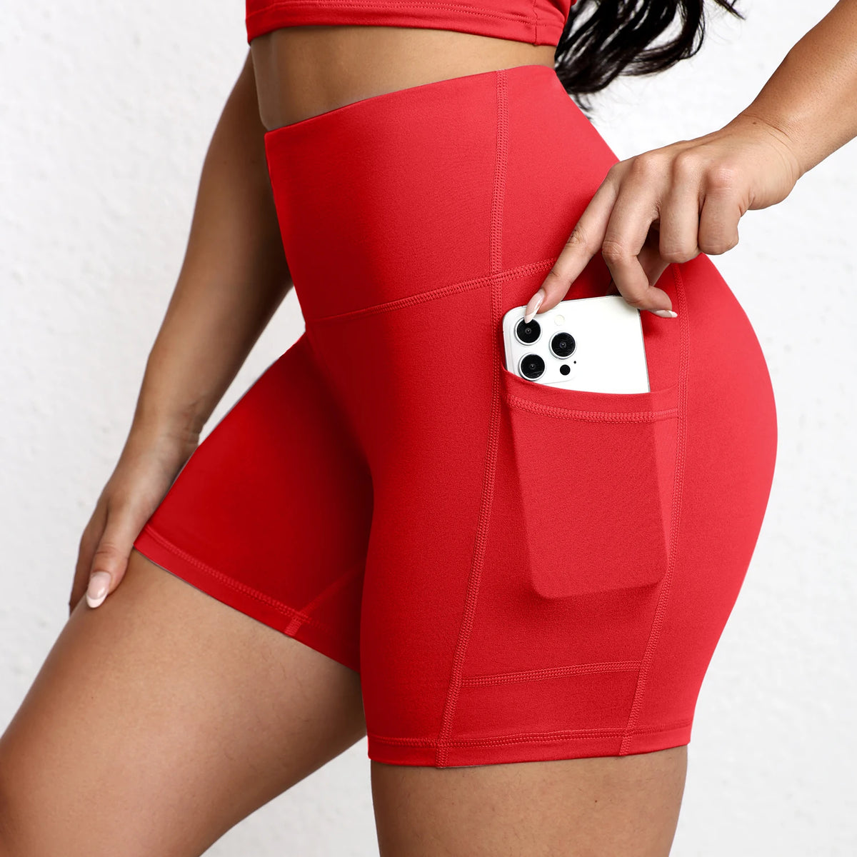 عارضة ترتدي شورت يوغا ورياضة أحمر وعلوي، وتظهر هاتفًا في الجيب.