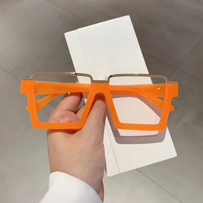 A hand holds a pair of bright orange oversized square semi-rim metal eyeglasses against a white background.