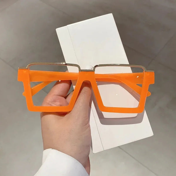 A hand holds a pair of bright orange oversized square semi-rim metal eyeglasses against a white background.