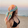 A woman wearing a silk-touch floral hair scarf in shades of peach and teal stands by the sea at sunset.