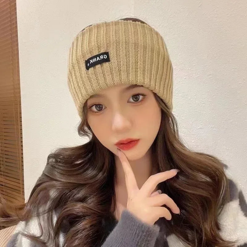 A woman wearing a beige knitted headband with black text on the front, looking directly at the camera.