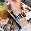 A hand wearing a leopard print silicone watch band is holding a glass of iced tea next to a menu.