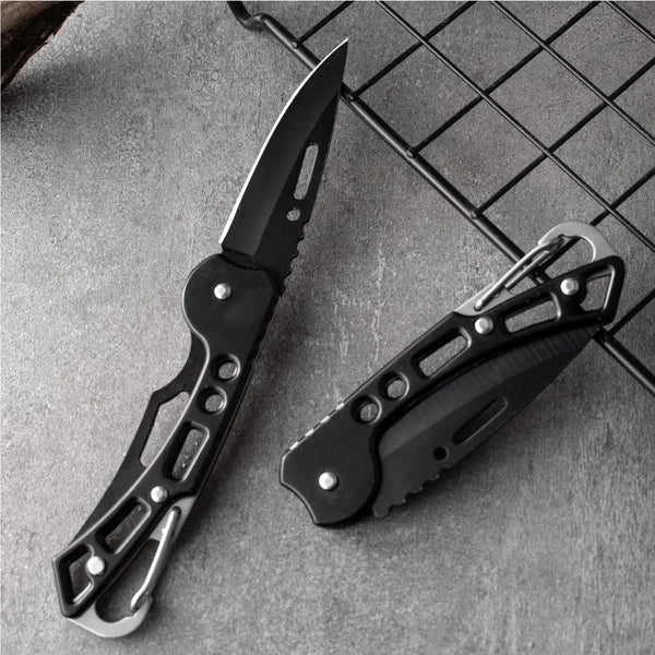 A black folding stainless steel survival knife is shown lying on a gray textured surface.