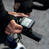 A hand is adjusting a black GPS cycling computer and speedometer mounted on a bicycle handlebar.