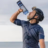 A man holds a blue and black protein shaker bottle up to his mouth outdoors.