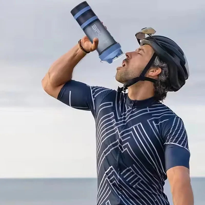 A man holds a blue and black protein shaker bottle up to his mouth outdoors.