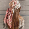 A woman shows the back of an elegant pink and cream satin square scarf tied in a flowing fabric.