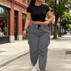 A woman wearing black crop top and gray drawstring cargo joggers is walking on a sidewalk.