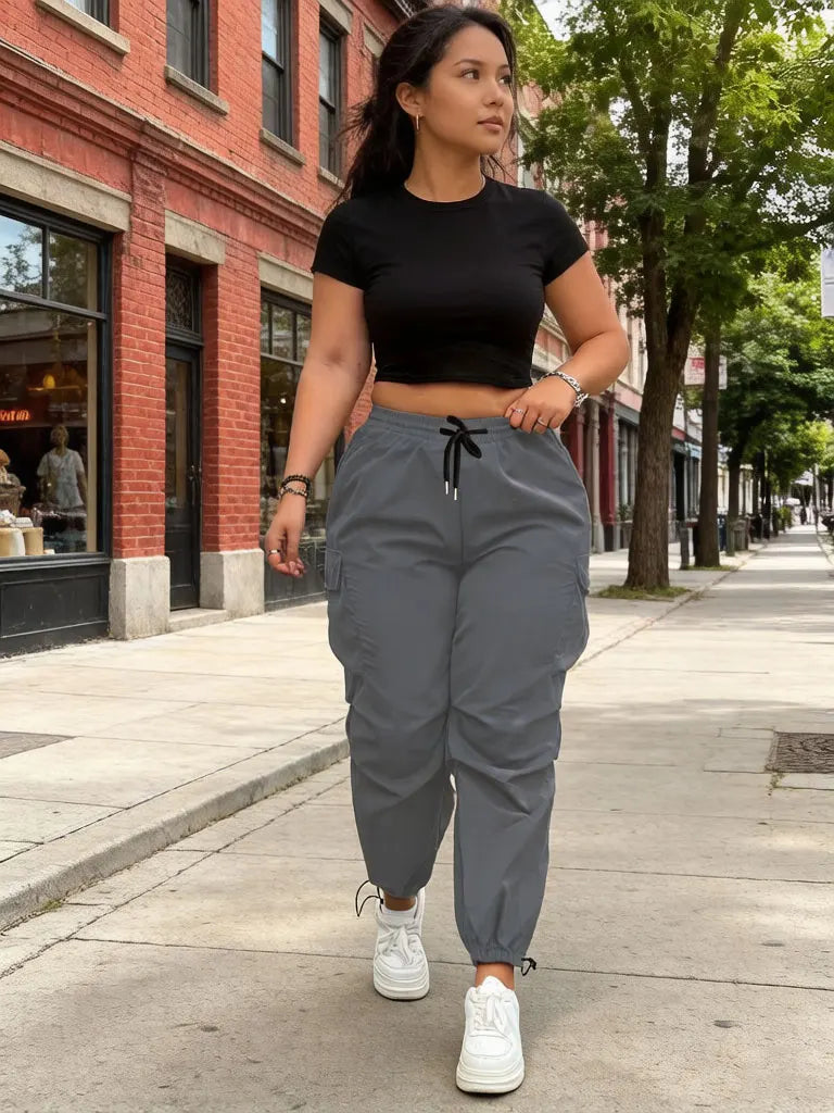 A woman wearing black crop top and gray drawstring cargo joggers is walking on a sidewalk.