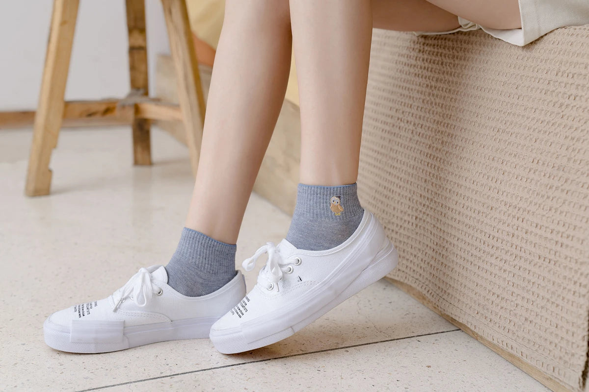 A pair of white canvas shoes with grey socks are shown resting on a light-colored floor next to a woven mat.