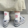 A pair of grey New Balance sneakers worn with white knitted socks and a light beige skirt.