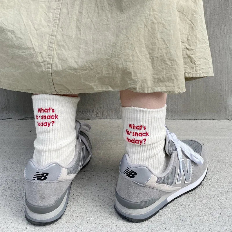 A pair of grey New Balance sneakers worn with white knitted socks and a light beige skirt.