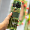 A hand holds a clear glass water bottle with a green cap and tea infuser, set against a blurred background of greenery.