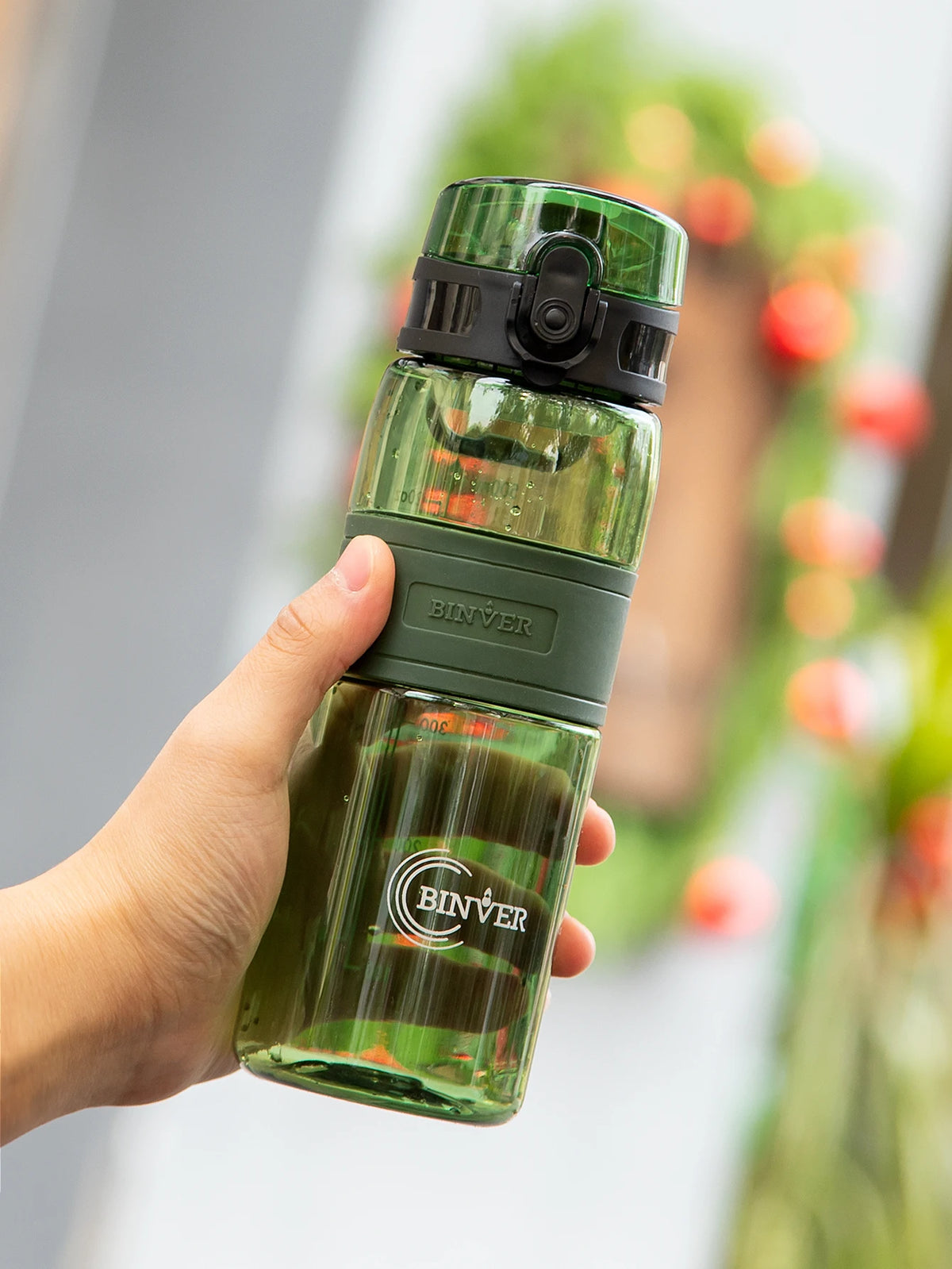 A hand holds a clear glass water bottle with a green cap and tea infuser, set against a blurred background of greenery.